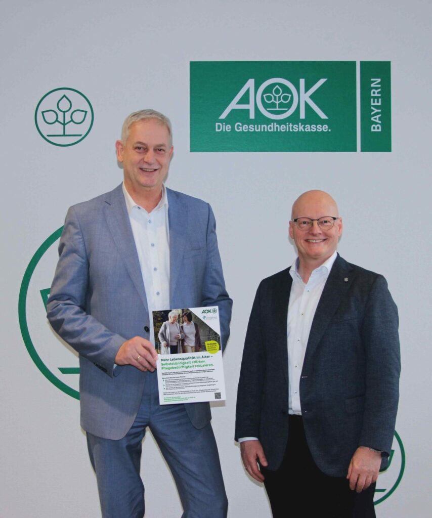 Zwei Männer in Anzügen stehen vor einem Schild der AOK Bayern, einer hält eine Broschüre.