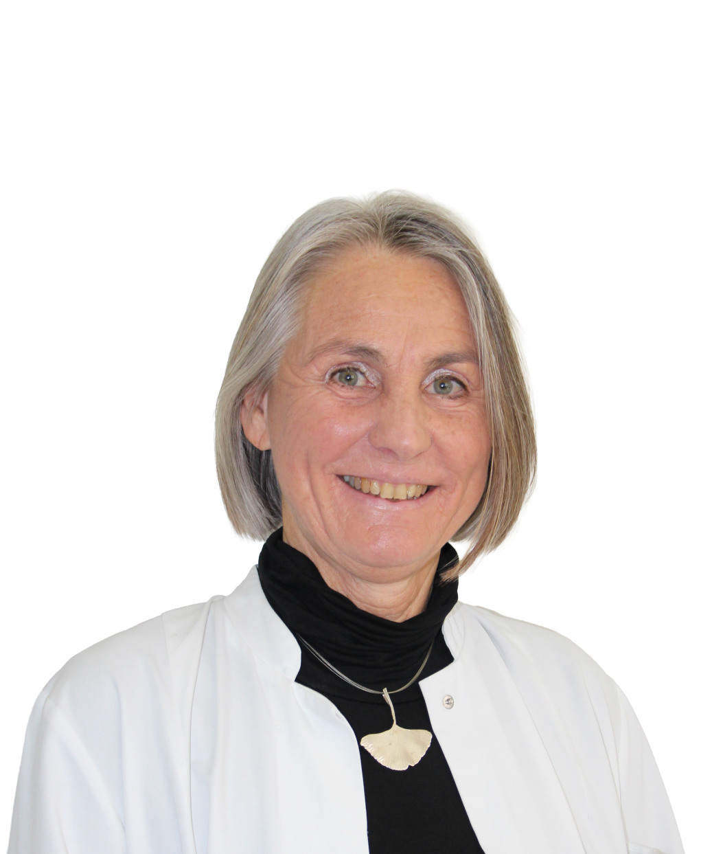 Eine Frau mit kurzen grauen Haaren, die einen weißen Mantel über einem schwarzen Rollkragenpullover und eine blattförmige Halskette mit Anhänger trägt, lächelt in die Kamera.